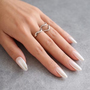 May include: A silver ring featuring a smooth, undulating wave design that wraps around the finger. The band has a polished, reflective metallic finish and a continuous, flowing shape that creates a subtle, curved aesthetic on the finger.