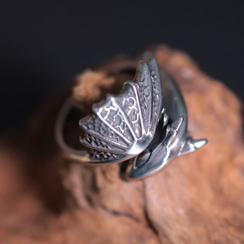 Op de afbeelding: Een zilverkleurige drakenring met gedetailleerde vleugel- en lichaamskenmerken. Het ontwerp van de ring omvat ingewikkelde patronen op de vleugels en een gebogen lichaam, waardoor een accessoire met fantasiethema ontstaat. De ring wordt getoond op een houten oppervlak.