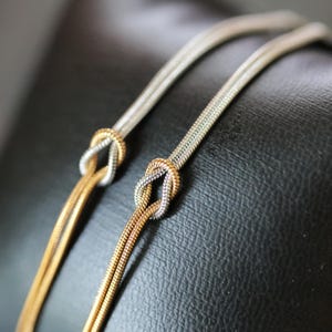 以下が含まれることがあります： 結び目デザインの蛇腹チェーンネックレス2本。1本はシルバーカラーのチェーンにゴールドカラーの結び目、もう1本はゴールドカラーのチェーンにシルバーカラーの結び目があしらわれています。チェーンは滑らかで柔軟な質感があり、光沢のある金属仕上げが施されています。