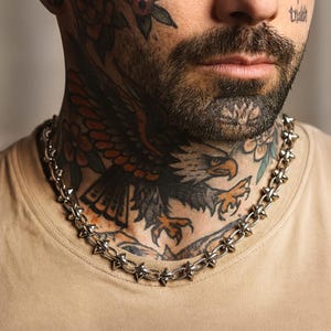 May include: A silver chain necklace featuring pointed spikes. The necklace is worn around the neck. The person in the image has tattoos on their neck and face. The background is a neutral colour.