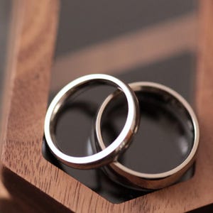 Könnte beinhalten: Zwei silberne Eheringe, einer in dem anderen verschachtelt, liegen auf einer dunklen Oberfläche in einer Holzbox. Die Ringe sind glatt und glänzend und reflektieren das Licht. Die Holzbox hat eine dunkelbraune Oberfläche.