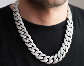 May include: A silver-colored, chunky chain necklace with a link design. The necklace is worn around the neck and rests on a black shirt. The chain is made of small, round, shiny pieces.