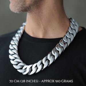 Puede incluir: Un collar de cadena plateada grande con un diseño de eslabones audaz y grueso. El collar mide aproximadamente 70 cm de largo y pesa unos 910 gramos. El collar se lleva sobre una camiseta negra.