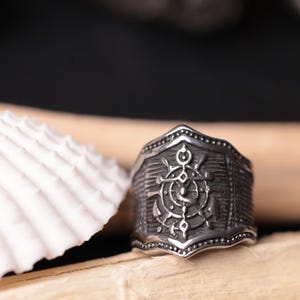Può includere: Un anello color argento con un design nautico raffigurante un'ancora e un timone. L'anello ha una fascia strutturata e una faccia dettagliata a forma di scudo. L'anello è esposto accanto a una conchiglia bianca.