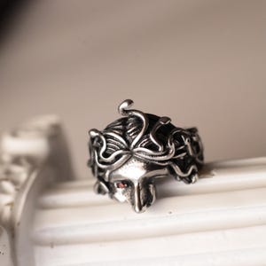 Könnte beinhalten: Ein silberfarbener Metallring mit einem skulptierten Gesicht und Details aus schlangenartigem Haar. Das Design enthält einen kleinen, runden roten Stein, der in die Augenhöhle des Gesichts eingesetzt ist und einen Kontrast zum Metall bildet.