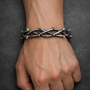 Può includere: Un bracciale in metallo scuro caratterizzato da un design continuo di spine intrecciate e appuntite. Il cinturino è composto da segmenti interconnessi che imitano l'aspetto di un tralcio spinoso, con una finitura in metallo grigio lucido.