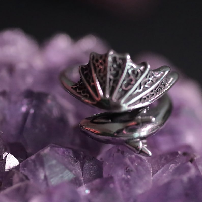Op de afbeelding: Een zilverkleurige ring met een drakenontwerp. De ring heeft een gedetailleerd, opengewerkt drakenvleugelontwerp en een drakenkop. De ring wordt getoond op een bed van paarse amethistkristallen.