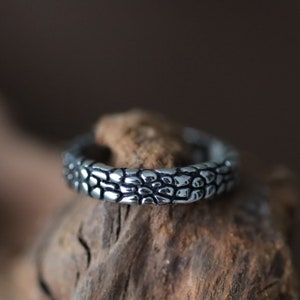 Könnte beinhalten: Ein silberfarbener Ring mit einem strukturierten Design, das an kleine Kieselsteine erinnert. Der Ring wird vor einem natürlichen Holzuntergrund präsentiert, der seinen metallischen Glanz hervorhebt. Die Oberfläche des Rings besteht aus unregelmäßigen Formen und erzeugt ein einzigartiges Muster.