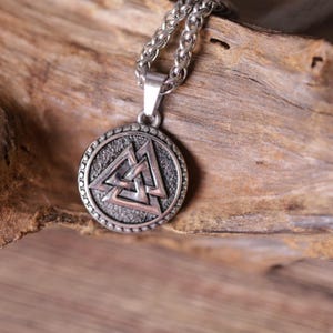 Puede incluir: Un collar con colgante plateado con un colgante circular. El colgante tiene un fondo texturizado y un diseño triangular central. El collar tiene una cadena con un patrón de eslabones texturizados. El colgante se muestra sobre un trozo de madera.