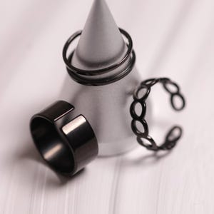 Könnte beinhalten: Drei schwarze Ringe in verschiedenen Designs werden auf einem grauen, kegelförmigen Ständer präsentiert. Ein Ring ist ein dickes Band mit offener Vorderseite, ein anderer hat ein Ketten-Design und der dritte hat zwei dünne Bänder.