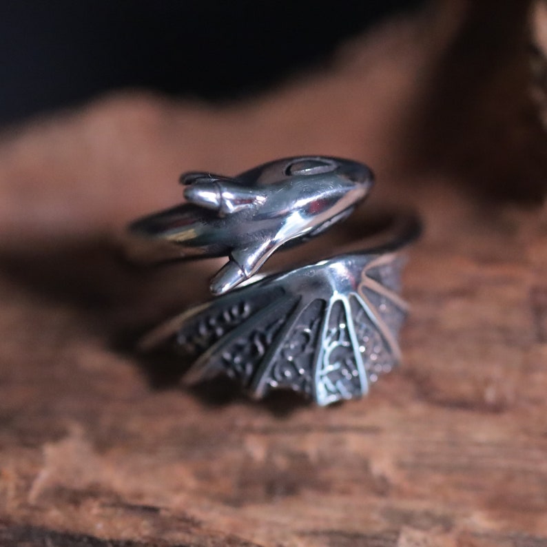 Op de afbeelding: Een zilveren drakenring. De ring heeft een drakenkop- en vleugelontwerp. De kop van de draak bevindt zich aan de bovenkant en de vleugels wikkelen zich om de vinger. De ring ligt op een houten oppervlak.