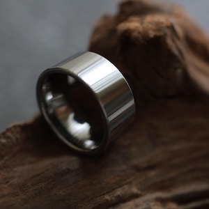 Op de afbeelding: Een brede, gepolijste zilveren ring met een glad, reflecterend oppervlak. De ring is geplaatst tegen een vage houten achtergrond, die de metaalglans benadrukt. Het ontwerp van de ring is eenvoudig en modern.