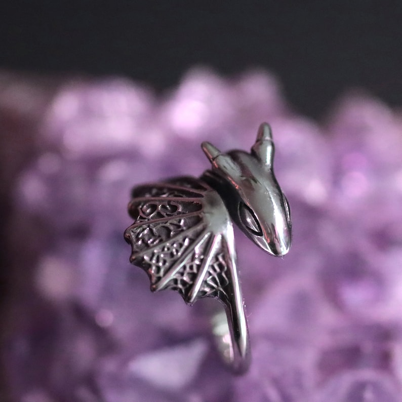 Op de afbeelding: Een zilverkleurige drakenring met gedetailleerde vleugels en een drakenkop. De ring is geplaatst tegen een wazige paarse achtergrond, die het ingewikkelde ontwerp van de sieraden benadrukt.