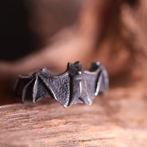 May include: A dark gray bat ring. The ring features a detailed bat design with outstretched wings. The ring is made of metal and has a textured surface. The ring is displayed on a wooden surface.