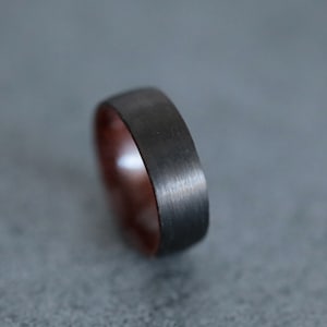 以下が含まれることがあります： つや消し金属仕上げの濃いグレーのリングのクローズアップ。リングの内側は温かみのある赤茶色です。リングはシンプルなバンドデザインです。