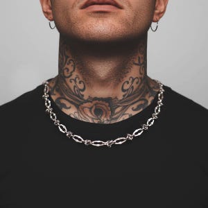 Könnte beinhalten: Silberfarbene Kette mit einem einzigartigen, strukturierten Gliederdesign. Die Halskette wird über einem schwarzen T-Shirt getragen, wodurch ein Kontrast entsteht, der den Schmuck hervorhebt. Die Kette ist ein auffälliges Accessoire.