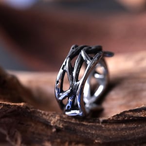 Peut inclure: Une bague argentée avec un design ouvert et entrelacé. La bague présente une série d'éléments incurvés en forme de branches qui créent un motif complexe. La bague est posée sur un fond en bois naturel, soulignant son éclat métallique.