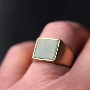May include: A gold-colored ring with a square, light green stone set in a gold bezel. The ring is on a finger, with a dark background. The ring's design is simple and elegant.