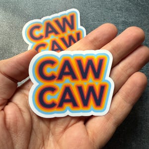 May include: Two stickers with the text "CAW CAW" in a retro font. The text is layered with orange, yellow, and blue outlines on a white background.