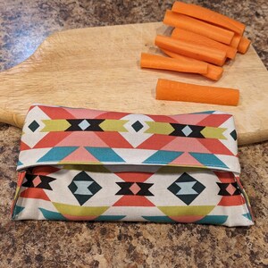May include: A colorful fabric food wrap with a geometric pattern in shades of pink, blue, green, and white. The wrap is folded in half and is lying on a wooden surface.