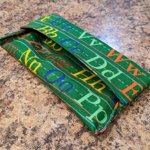 May include: Green fabric pencil case with a repeating pattern of letters and dashed lines. The case is folded closed.