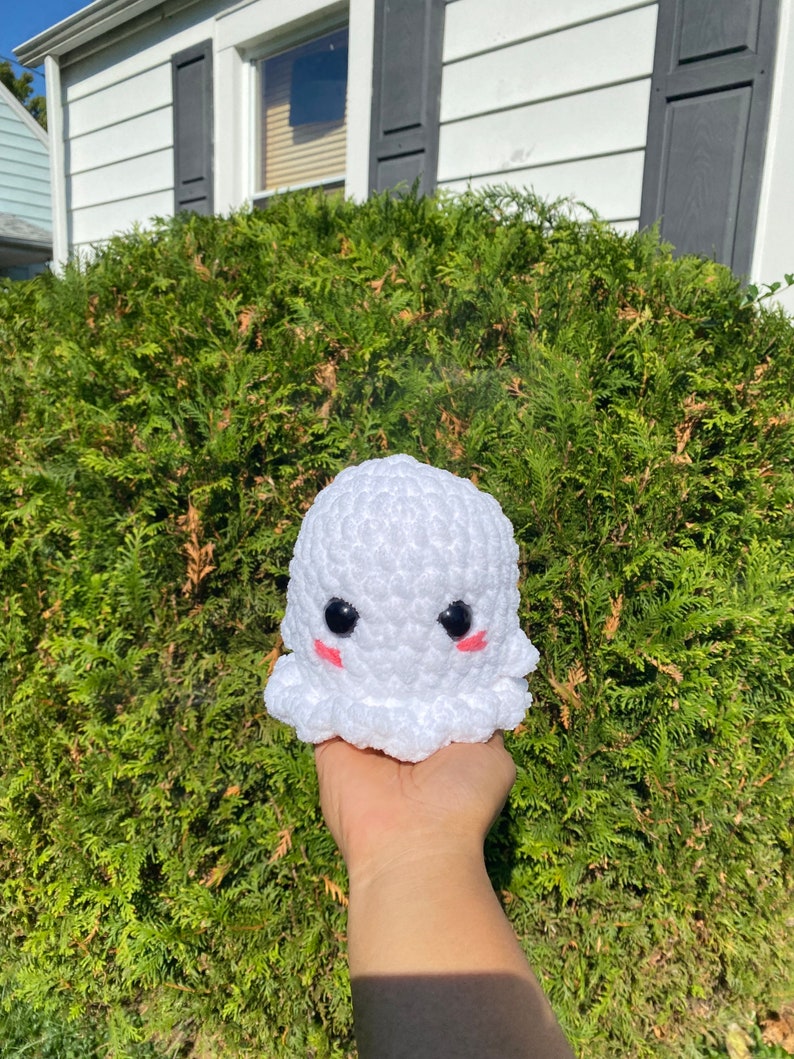 May include: A white crocheted ghost plush toy with black eyes and pink cheeks. The ghost is being held in a person's hand.
