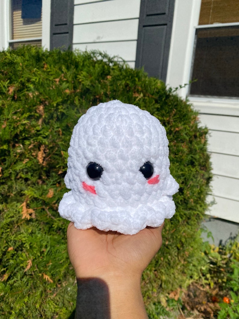 May include: A white crocheted ghost with black eyes and pink cheeks. The ghost is holding a small white crocheted heart.