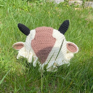 May include: A white and brown crocheted hat shaped like a cow with black ears. The hat is sitting on a bed of green grass.