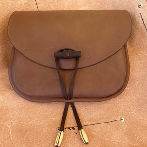 May include: A brown leather bag with a flap closure and a drawstring closure. The bag has a brown leather strap with a metal buckle.