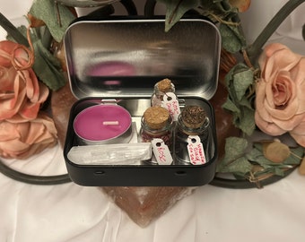 May include: A black metal tin containing a pink candle, a selenite wand, and small glass jars with cork stoppers. The jars are labeled "Rose Quartz", "Rose", and "Hawaiian Black Sea Salt". The tin is surrounded by pink roses and decorative elements.