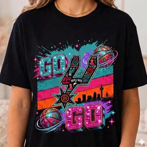 Może przedstawiać: Czarny t-shirt z nadrukiem graficznym z napisem GO GO. Projekt zawiera stylizowane logo Spurs, piłki do koszyk&oacute;wki z motywami westernowymi, sylwetkę panoramy miasta oraz kolorowe poziome paski w odcieniach turkusu, r&oacute;żu i pomarańczu.
