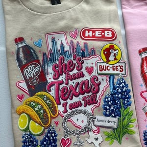 Può includere: Una t-shirt beige con una stampa grafica che riporta il testo 'she's from Texas I can tell'. Il design include una bottiglia di Dr Pepper, tacos, lime, lupini, un braccialetto con ciondoli e i loghi H-E-B e Buc-ee's su una mappa del Texas.