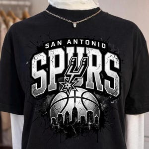 Może przedstawiać: Czarny t-shirt z kr&oacute;tkim rękawem z nadrukiem graficznym przedstawiającym napis SAN ANTONIO SPURS białymi literami. Wz&oacute;r zawiera piłkę do koszyk&oacute;wki, logo Spurs oraz sylwetkę panoramy miasta z efektem popękanej tekstury.