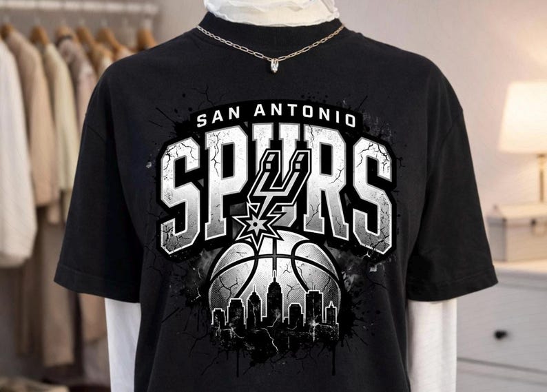 Może przedstawiać: Czarny t-shirt z kr&oacute;tkim rękawem z nadrukiem graficznym przedstawiającym napis SAN ANTONIO SPURS białymi literami. Wz&oacute;r zawiera piłkę do koszyk&oacute;wki, logo Spurs oraz sylwetkę panoramy miasta z efektem popękanej tekstury.