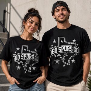 Może przedstawiać: Czarny t-shirt z kr&oacute;tkim rękawem z białą grafiką stanu Teksas, napisem "GO SPURS GO" oraz kilkoma gwiazdkami. Projekt zawiera małe logo związane z koszyk&oacute;wką wewnątrz konturu stanu. Widoczny jest r&oacute;wnież napis "DIGITAL DOWNLOAD".