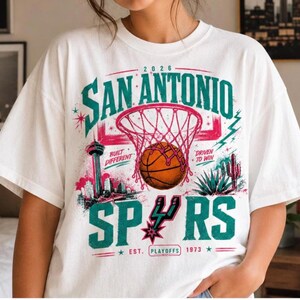 Może przedstawiać: Biały t-shirt z kr&oacute;tkim rękawem z nadrukiem graficznym zawierającym tekst San Antonio, 2026, Built Different, Driven To Win, Spurs oraz Est. Playoffs 1973. Wz&oacute;r przedstawia piłkę do koszyk&oacute;wki, kosz, panoramę miasta i ilustrację kaktusa.