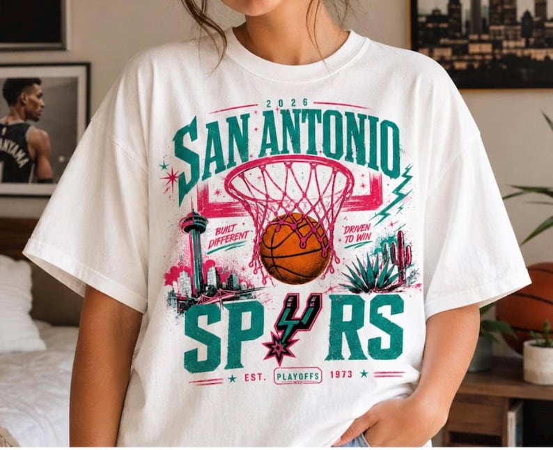 Może przedstawiać: Biały t-shirt z kr&oacute;tkim rękawem z nadrukiem graficznym zawierającym tekst San Antonio, 2026, Built Different, Driven To Win, Spurs oraz Est. Playoffs 1973. Wz&oacute;r przedstawia piłkę do koszyk&oacute;wki, kosz, panoramę miasta i ilustrację kaktusa.