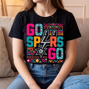 Może przedstawiać: Czarny t-shirt z kr&oacute;tkim rękawem z kolorowym nadrukiem graficznym z napisem GO SPURS GO pogrubionymi, blokowymi literami. Projekt zawiera centralne logo gwiazdy oraz geometryczne wzory w żywych odcieniach r&oacute;żu, turkusu, pomarańczu i ż&oacute;łci.