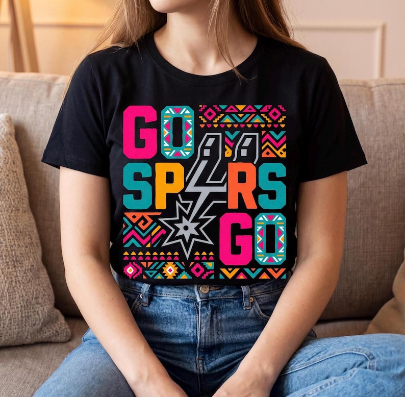 Może przedstawiać: Czarny t-shirt z kr&oacute;tkim rękawem z kolorowym nadrukiem graficznym z napisem GO SPURS GO pogrubionymi, blokowymi literami. Projekt zawiera centralne logo gwiazdy oraz geometryczne wzory w żywych odcieniach r&oacute;żu, turkusu, pomarańczu i ż&oacute;łci.