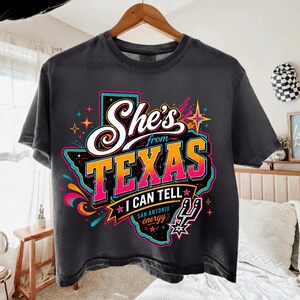 Może przedstawiać: Ciemnoszary t-shirt z grafiką stanu Teksas w kolorze czarnym z jasnym neonowym konturem. Napis brzmi "She's from TEXAS I CAN TELL" białymi i pomarańczowymi literami z gradientem, z dopiskiem "SAN ANTONIO energy" oraz logo koszyk&oacute;wki.