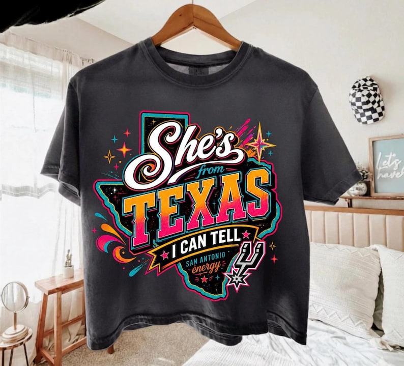 Może przedstawiać: Ciemnoszary t-shirt z grafiką stanu Teksas w kolorze czarnym z jasnym neonowym konturem. Napis brzmi "She's from TEXAS I CAN TELL" białymi i pomarańczowymi literami z gradientem, z dopiskiem "SAN ANTONIO energy" oraz logo koszyk&oacute;wki.