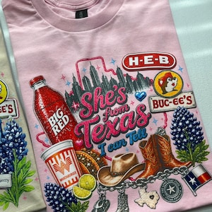 Può includere: Una t-shirt rosa chiaro con una stampa grafica che riporta il testo "She's from Texas I can Tell". Il design include una bottiglia di soda Big Red, un bicchiere Whataburger, un cappello da cowboy, stivali, fiori e icone a tema Texas.