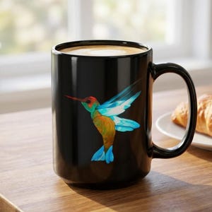 May include: A black ceramic mug featuring a vibrant, multi-colored hummingbird illustration on the front. The hummingbird displays shades of blue, orange, and red with a long, thin beak. The mug has a smooth, glossy finish and a curved handle.