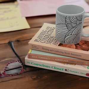 May include: A stack of three books with a white mug with a black wavy pattern on top. The top book is titled "The Conscious Closet" by Elizabeth L. Cline. The middle book is titled "Toxic Femininity in the Workplace" by Ginny Hogan. The bottom book is titled "Planning and Conducting Needs Assessments" by Witkin Altschuld.