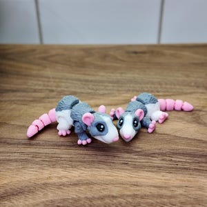 May include: Two handmade opossum figurines. The opossums are gray and white with pink ears, feet, and tails. They are lying on a wooden surface. The figurines are detailed and whimsical.