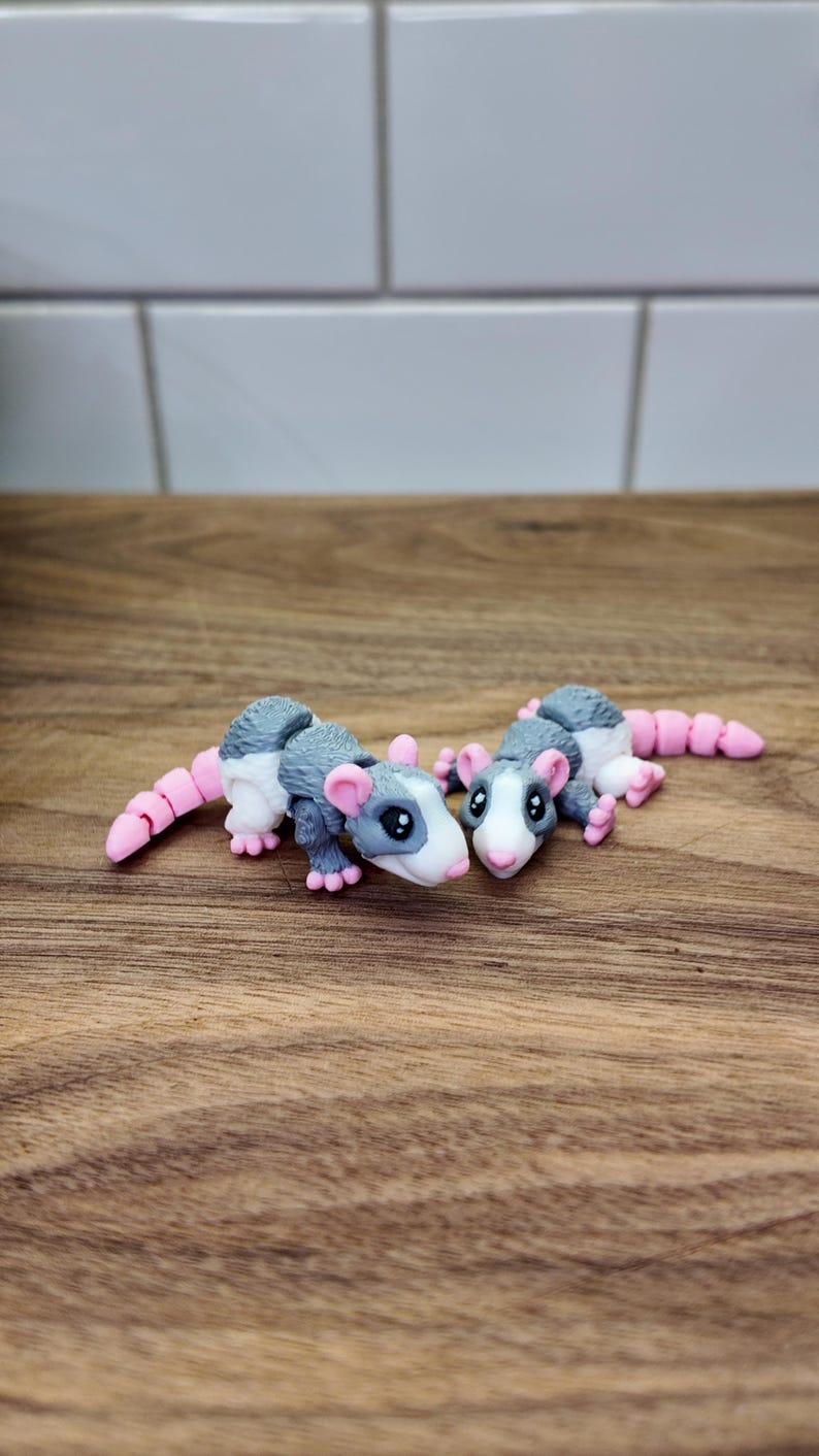 May include: Two handmade opossum figurines. The opossums are gray and white with pink ears, feet, and tails. They are lying on a wooden surface. The figurines are detailed and whimsical.
