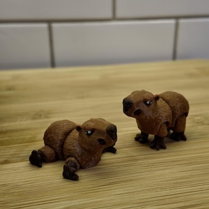 May include: Two brown capybara figurines, one lying down and the other standing. The figurines are detailed with textured fur and small black feet. They are set on a light wood surface against a white tile background.