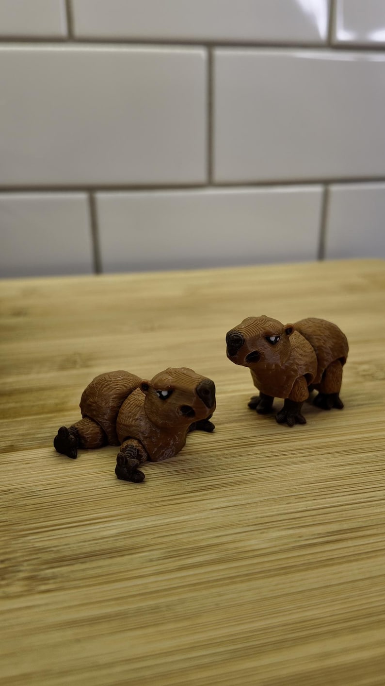 May include: Two brown capybara figurines, one lying down and the other standing. The figurines are detailed with textured fur and small black feet. They are set on a light wood surface against a white tile background.