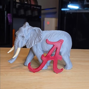 Pode incluir: Uma estatueta de elefante cinzento com presas de marfim está ao lado de uma letra "A" vermelha. O elefante é detalhado com rugas e dobras, e a letra está em uma fonte arrojada e estilizada. Os objetos estão em uma superfície castanho-clara.