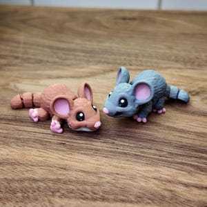 May include: Two small, detailed mouse figurines. One is brown with pink accents, the other is gray with pink accents. Both have large eyes and are lying on a wooden surface.